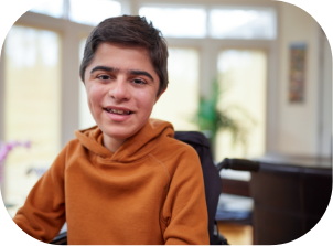 child in a wheelchair wearing an orange hoodie, smiling inside a bright living room with windows.