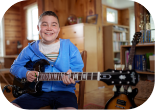 child smiling while playing a black electric guitar inside a cozy room with wood paneling.