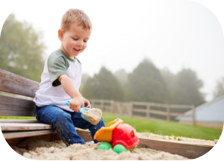 Young child playing in a sandbox with a toy truck on a foggy day near a wooden fence.