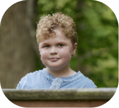 Portrait of a young child with curly blond hair wearing a blue shirt, outdoors with trees in the background.